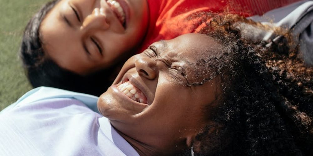 High angle of cheerful multiracial girlfriends lying on green lawn and toothy smiling while having fun together